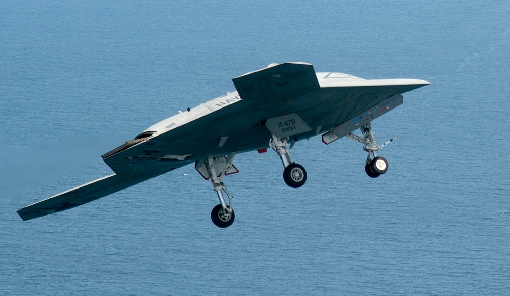 un X-47B en vol au-dessus de la mer, vu de profil droit du dessous, trains d'd’atterrissage sortis