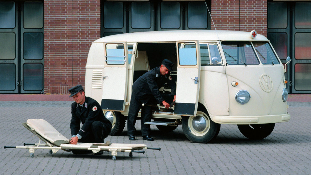 vu de 3/4 avant gauche, double portes latérales ouvertes, 1 ambulancier prépare une civière, l'autre s'occupe de quelque chose près du véhicule, un marche-pied a été ajouté au T1