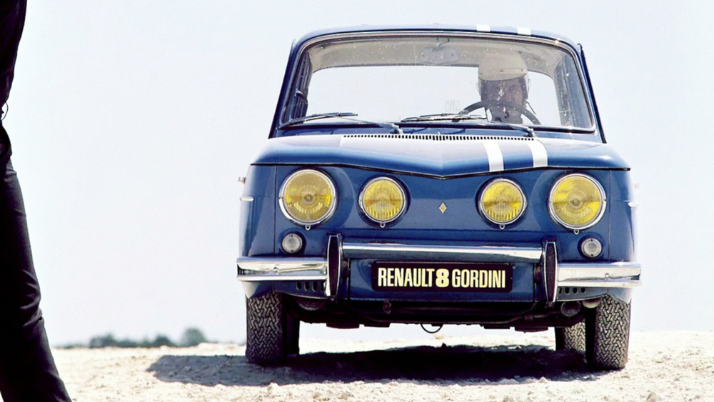 une Renault 8 Gordini 1964–1970 vue de face, un homme sur le côté gauche de la photo agite un drapeau à damier